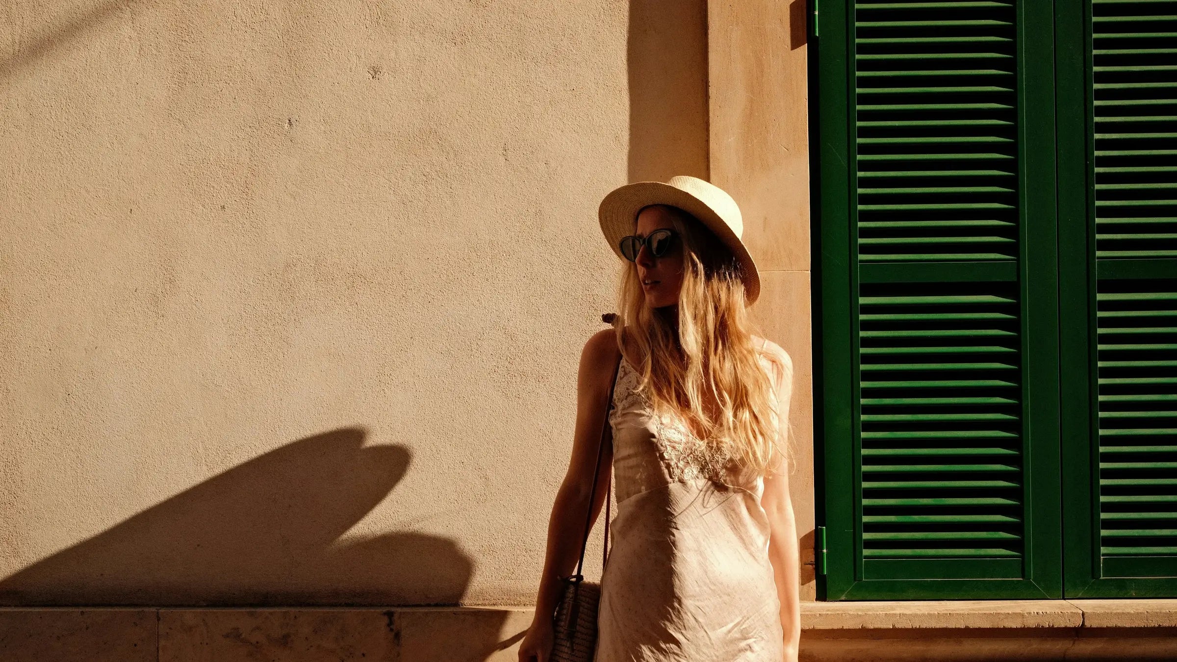 Mujer con vestido lencero y sombrero de paja junto a una contraventana verde