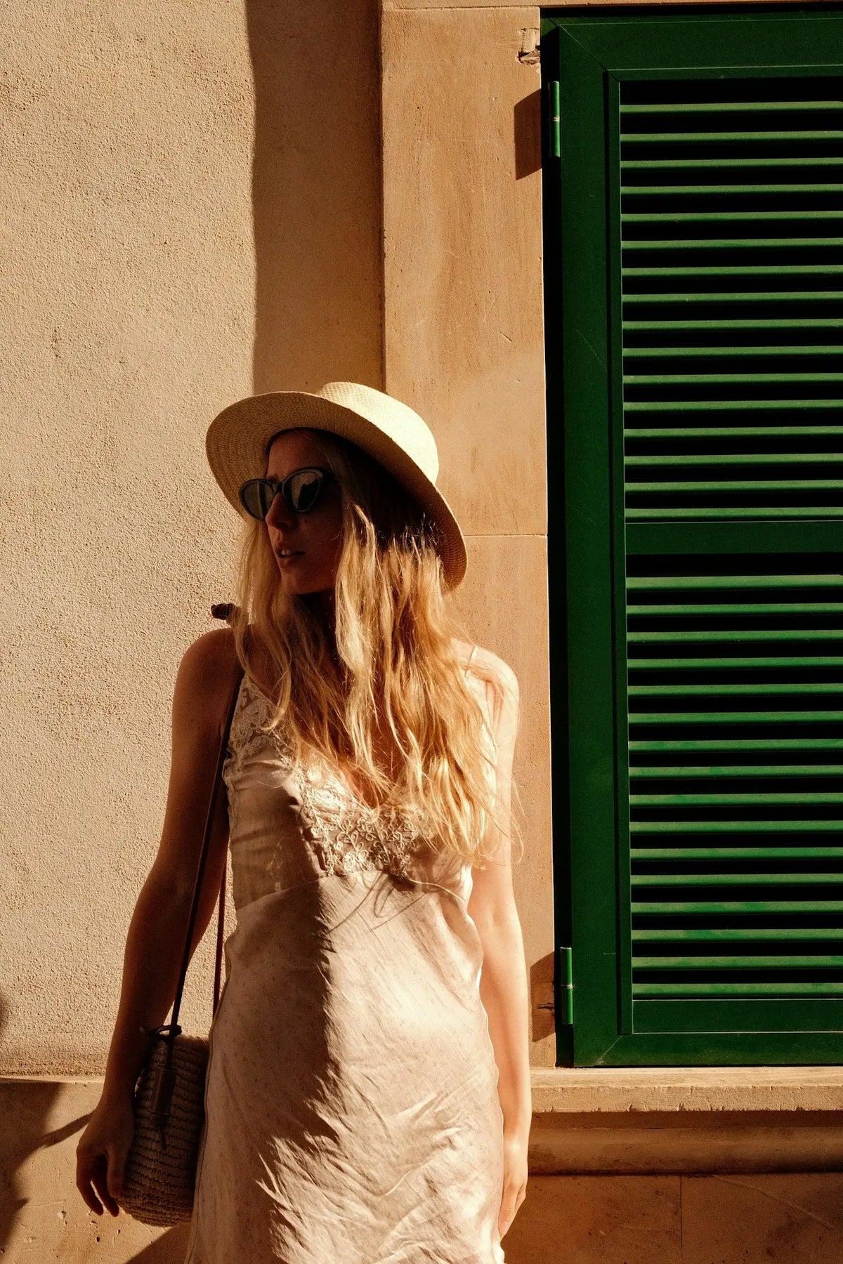 Mujer con vestido lencero y sombrero de paja junto a una contraventana verde
